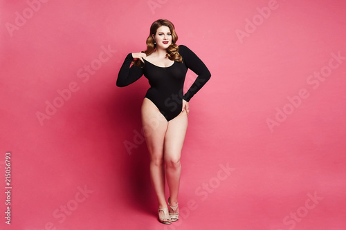 Plus size sexy model girl, fashionable blonde in black bodysuit, with bright makeup and with stylish hairstyle, smiling and posing at pink background in studio