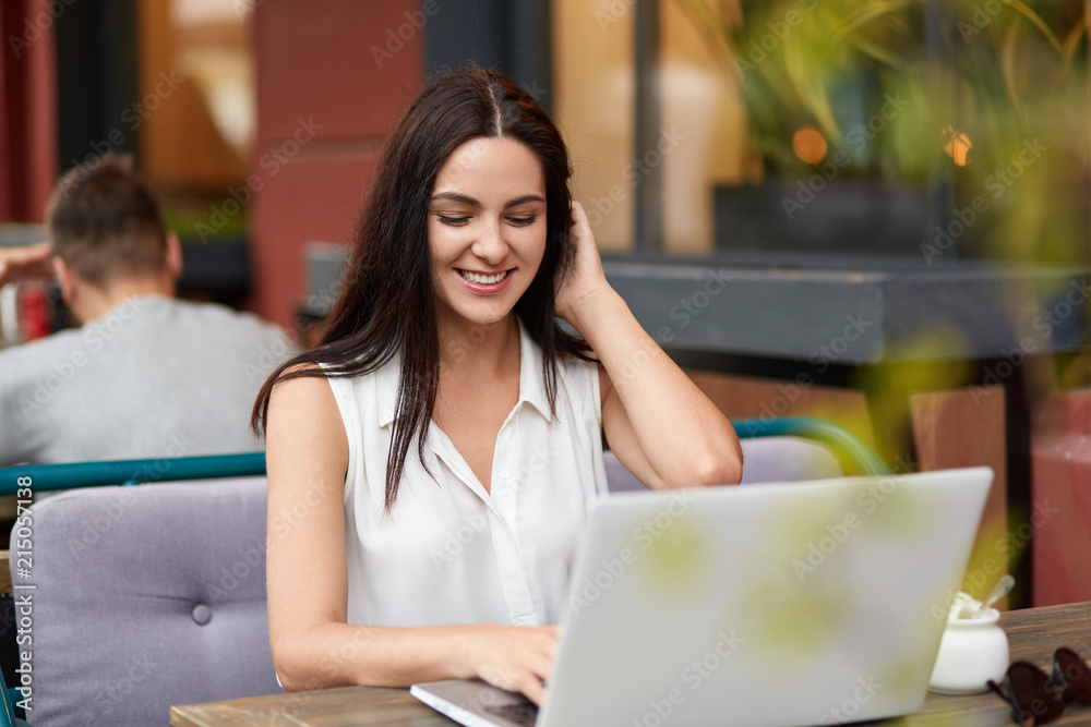 Cheerful young female freelancer works on laptop computer in bistro, has toothy smile as finishes work, dressed in white blouse, browses internet webpage connected to wireless wifi. Technology concept Cheerful young female freelancer works on laptop computer in bistro, has toothy smile as finishes work, dressed in white blouse, browses internet webpage connected to wireless wifi. Technology concept