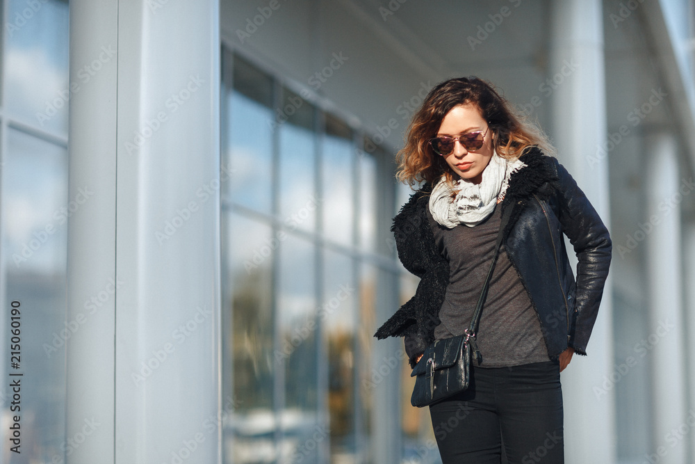 woman in sun glasses a black leather jacket, black jeans posing in front of mirrored windows. Female fashion concept. Outdoor
