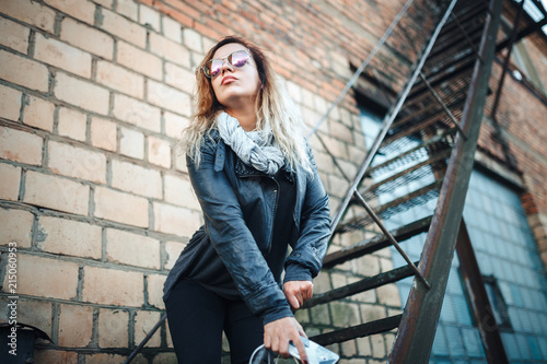 Smiling young woman in sunglasses, a black leather jacket, black jeans standing on an urban metal stair against a brick wall and dances. Woman listening to music on the stairs of industrial building.