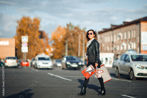 Lovely young woman in sunglasses  a black leather jacket  black jeans turns around holding shopping bags on Parking near the shopping center.