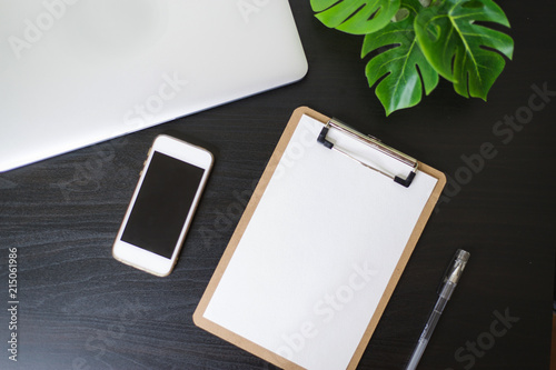 Still life, business, office supplies or education concept : Top view image of open notebook, clip board with blank pages, laptope, pen, phone and flower on wooden dark background, ready for adding or photo