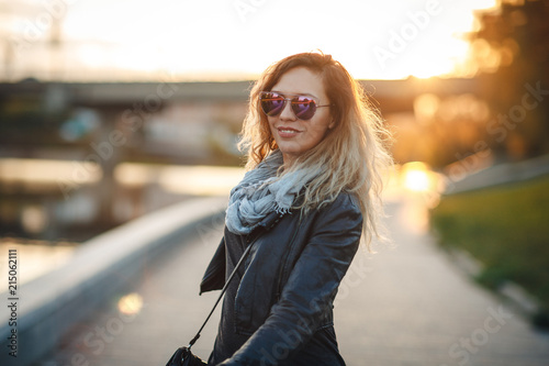 Attractive woman in mirrored sunglasses, a black leather jacket, black jeans walking along the river Bank in the city, turns to camera and smiles in front the setting sun. Waving hair. city,bridge