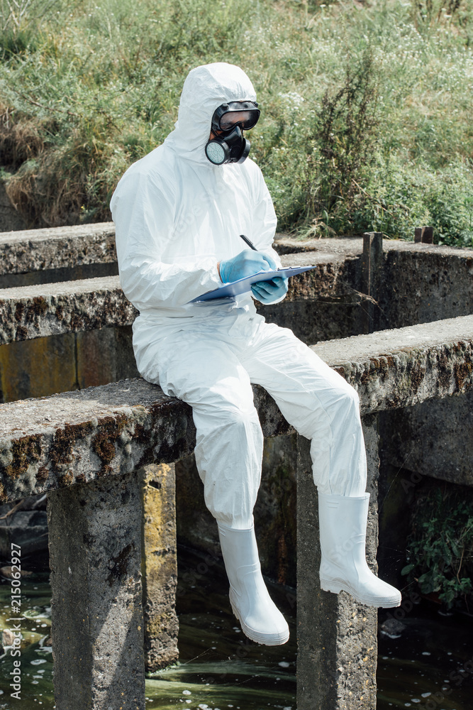 selective focus of male scientist in protective mask and suit writing in clipboard while sitting near sewerage