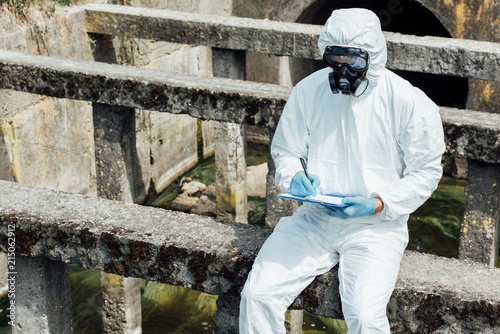 Slika na platnu male scientist in protective mask and suit writing in clipboard while sitting n