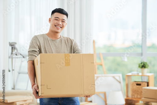 Fotografie Happy cheerful ethnic male in casual clothes carrying cardboard box and looking