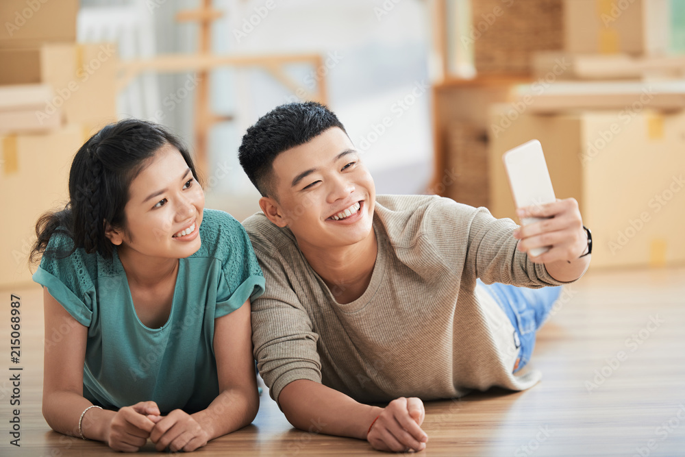 Happy young Asian couple lying on floor of new apartment on moving day and taking picture on smartphone front camera smiling on blurred background 