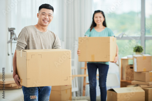 Cheerful young Asian man and woman holding large cardboard boxes for moving and looking at camera smiling on blurred background  © DragonImages