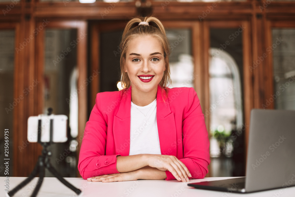 Beautiful smiling girl in pink jacket happily looking in camera while recording new video with cellphone and laptop Beautiful smiling girl in pink jacket happily looking in camera while recording new video with cellphone and laptop