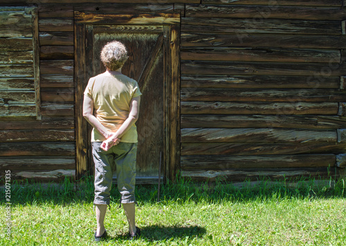 rear view of middle aged Caucasian woman standing in the grass looking at the closed wooden door of a weathered antique barn with copy space