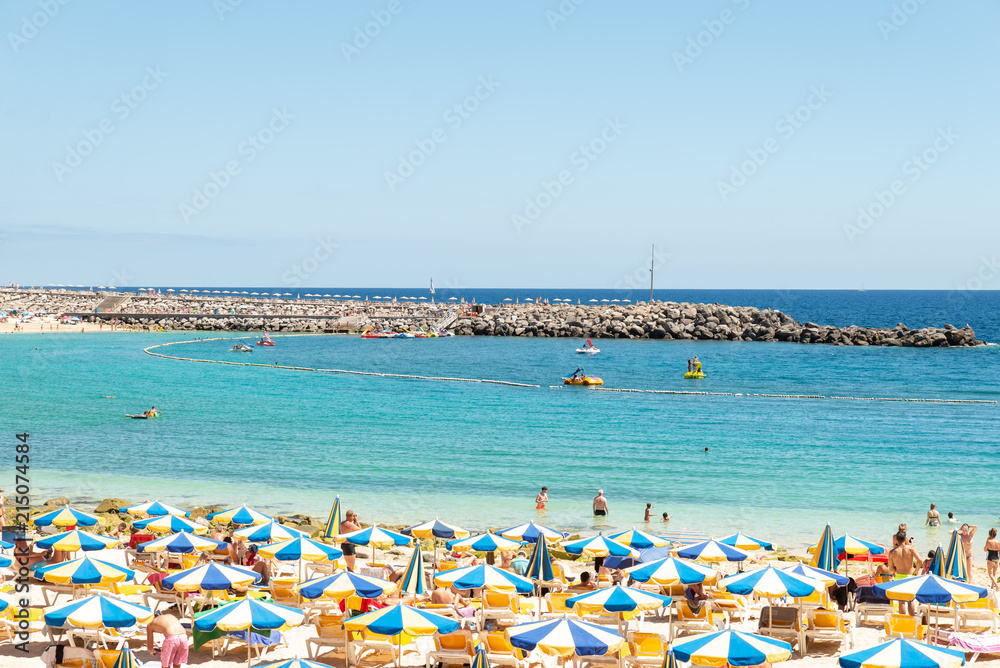 Las Americas, Canarias islands/ Spain-July 22, 2018: View of beach fool of tourists and umbrellas for sun on Canarias, view of bay with mountains. Blue Atlantic ocean and beach on Gran Canaria. Las Americas, Canarias islands/ Spain-July 22, 2018: View of beach fool of tourists and umbrellas for sun on Canarias, view of bay with mountains. Blue Atlantic ocean and beach on Gran Canaria.