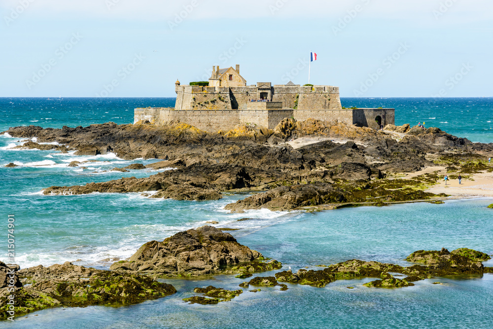 The Fort National built by french military architect Vauban on a tidal island, seen from the city of Saint-Malo, France, with the french flag blowing in the wind and rocks in the sea in the foreground