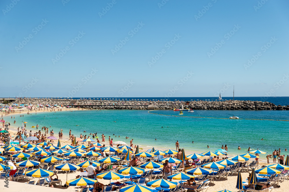 Las Americas, Canarias islands/ Spain-July 22, 2018: View of beach fool of tourists and umbrellas for sun on Canarias, view of bay with mountains. Blue Atlantic ocean and beach on Gran Canaria. Las Americas, Canarias islands/ Spain-July 22, 2018: View of beach fool of tourists and umbrellas for sun on Canarias, view of bay with mountains. Blue Atlantic ocean and beach on Gran Canaria.