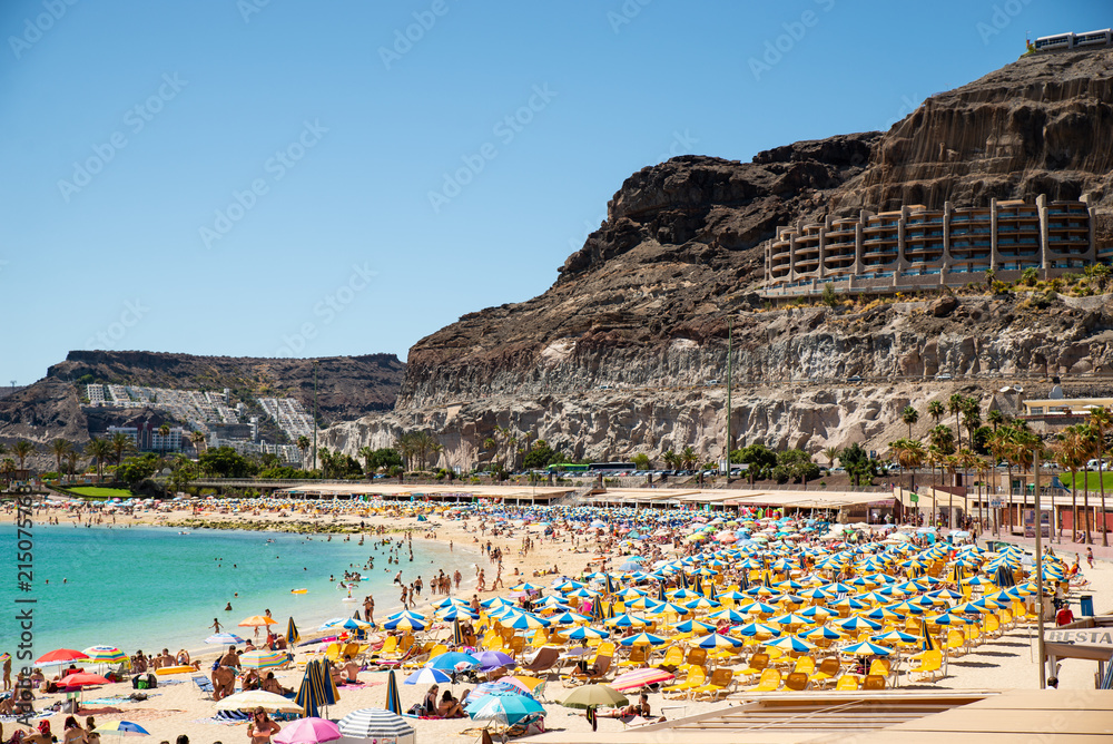 Las Americas, Canarias islands/ Spain-July 22, 2018: View of beach fool of tourists and umbrellas for sun on Canarias, view of bay with mountains. Blue Atlantic ocean and beach on Gran Canaria. Las Americas, Canarias islands/ Spain-July 22, 2018: View of beach fool of tourists and umbrellas for sun on Canarias, view of bay with mountains. Blue Atlantic ocean and beach on Gran Canaria.