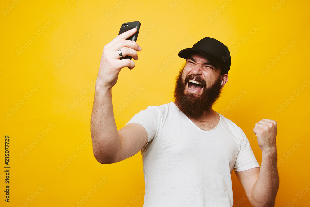 Portrait of a happy young bearded man in white t-shirt looking at mobile phone and celebrating isolated over yellow background Portrait of a happy young bearded man in white t-shirt looking at mobile phone and celebrating isolated over yellow background