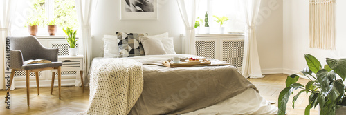 Real photo of bright bedroom interior with king-size bed with wooden tray with breakfast and knit blanket, fresh green plants and grey armchair with book