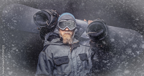 Fototapeta Naklejka Na Ścianę i Meble -  Brutal redhead snowboarder with a full beard in a winter hat and protective glasses dressed in a snowboarding coat posing with snowboard at a studio.