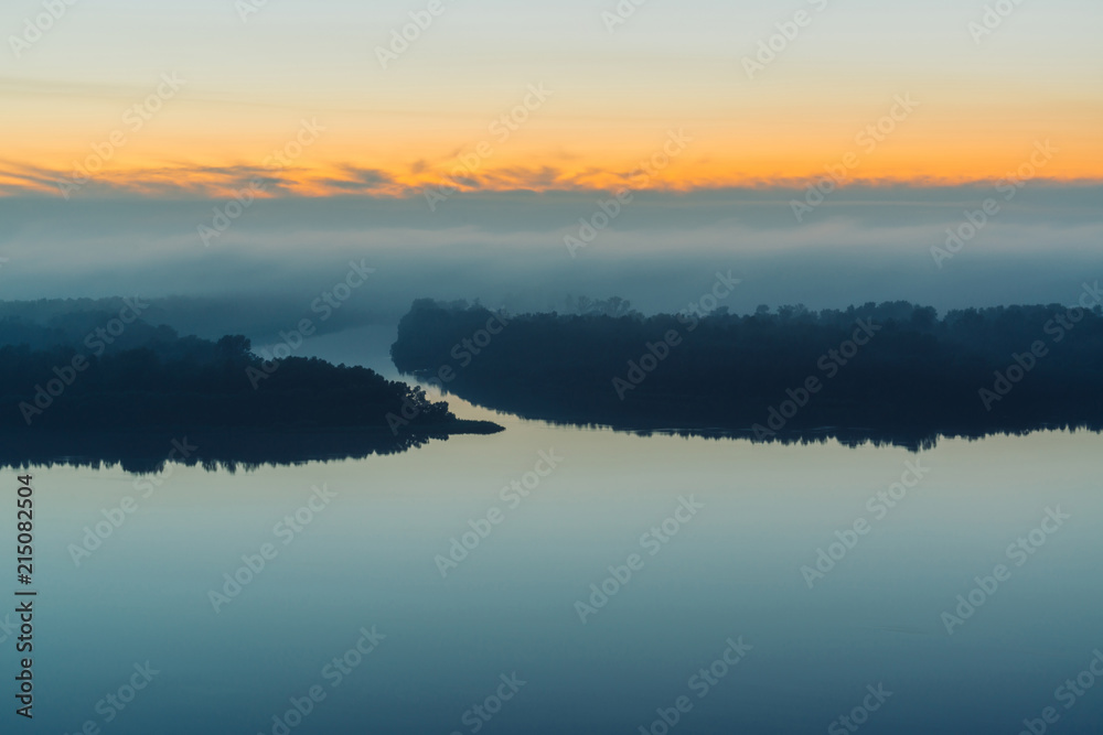 Broad river flows along shore with forest under thick fog. Early blue sky reflected in water. Yellow glow in picturesque predawn sky. Colorful morning mystical atmospheric landscape of majestic nature
