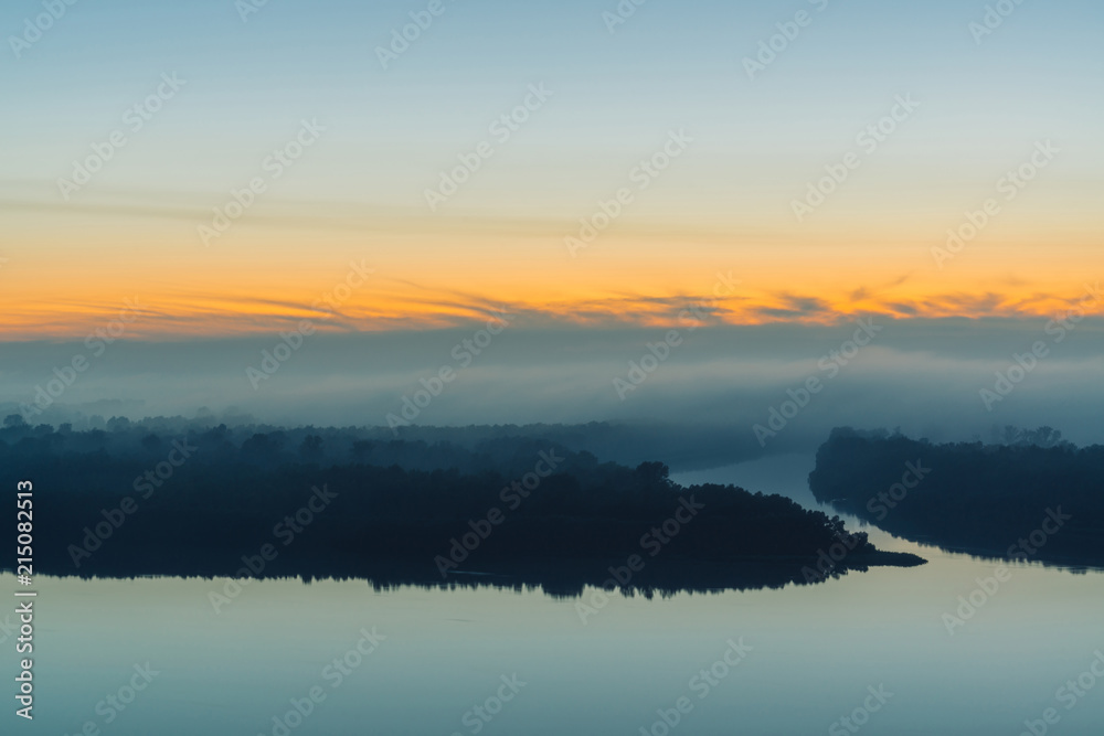 Broad river flows along shore with forest under thick fog. Early blue sky reflected in water. Yellow glow in picturesque predawn sky. Colorful morning mystical atmospheric landscape of majestic nature