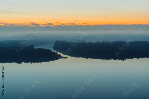 Fototapeta Naklejka Na Ścianę i Meble -  Broad river flows along shore with forest under thick fog. Early blue sky reflected in water. Yellow glow in picturesque predawn sky. Colorful morning mystical atmospheric landscape of majestic nature
