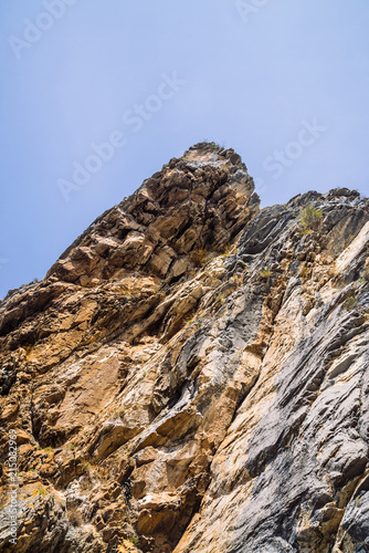 Big mountain cliff under cloudy sky close-up. Beautiful rocky gray textured background with vegetation. Majestic nature.