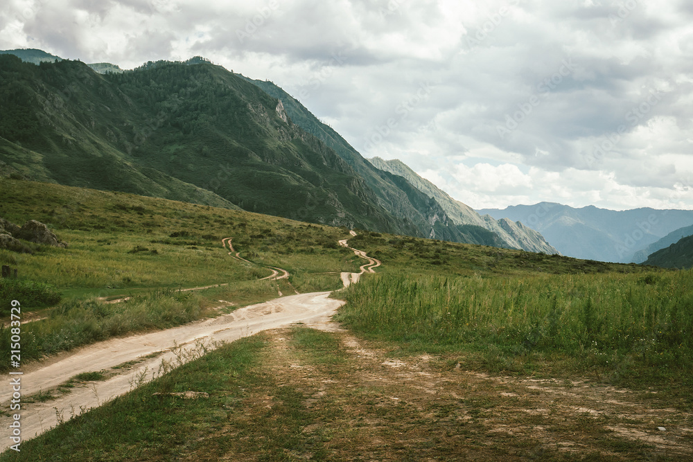 Dirt road leads to mountains in highland. Green grass near road. Beautiful mountain terrain under cloudy sky. Overcast weather in wild. Atmospheric tranquil landscape of majestic nature.