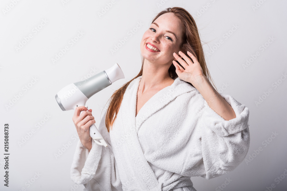 Beautiful red-haired girl in a white bathrobe bathing her hair with a hair dryer on a light background Beautiful red-haired girl in a white bathrobe bathing her hair with a hair dryer on a light background