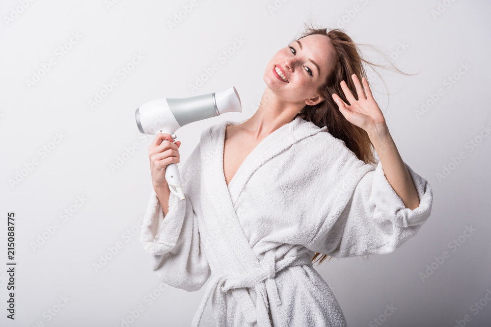Beautiful red-haired girl in a white bathrobe bathing her hair with a hair dryer on a light background Beautiful red-haired girl in a white bathrobe bathing her hair with a hair dryer on a light background