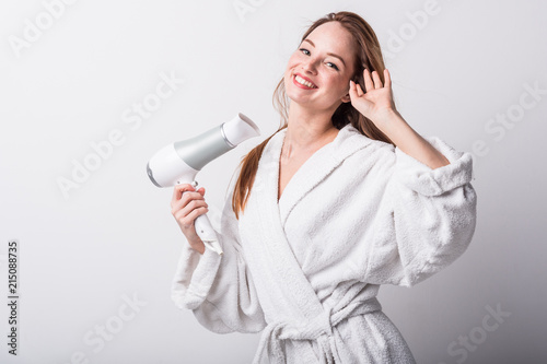  Beautiful red-haired girl in a white bathrobe bathing her hair with a hair dryer on a light background