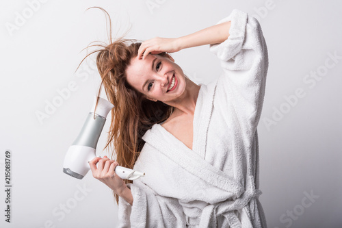  Beautiful red-haired girl in a white bathrobe bathing her hair with a hair dryer on a light background
