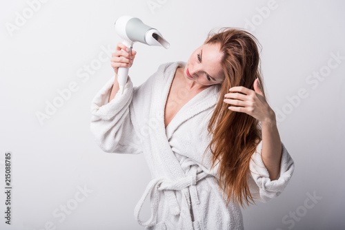  Beautiful red-haired girl in a white bathrobe bathing her hair with a hair dryer on a light background
