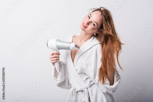  Beautiful red-haired girl in a white bathrobe bathing her hair with a hair dryer on a light background