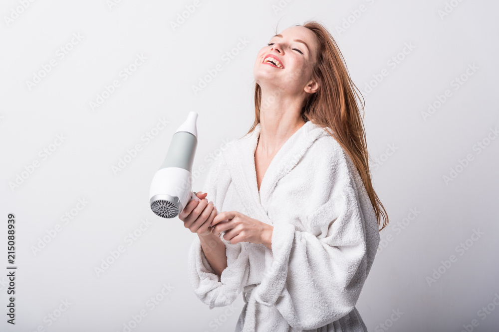 Beautiful red-haired girl in a white bathrobe bathing her hair with a hair dryer on a light background Beautiful red-haired girl in a white bathrobe bathing her hair with a hair dryer on a light background