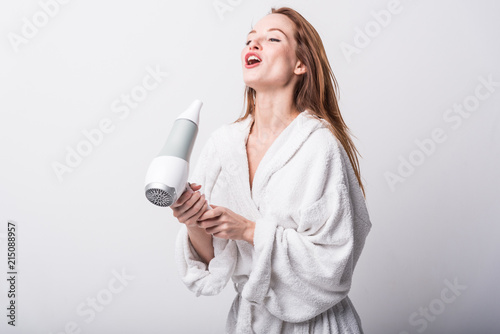  Beautiful red-haired girl in a white bathrobe bathing her hair with a hair dryer on a light background