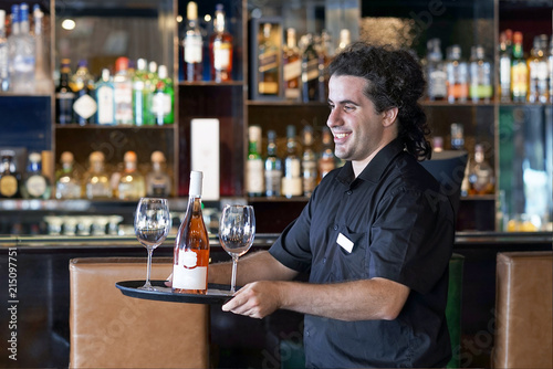 A young men bartender carries a bottle of red wine with wine glasses on a tray to the client of the hote bar, restaurant.  The concept of service.