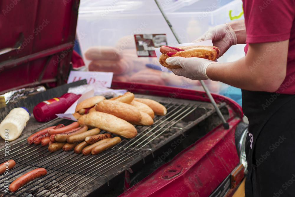 A man is cooking hot dogs in a barbeque car on the street.