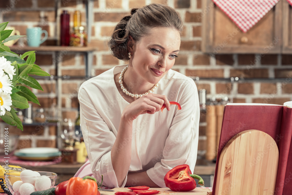 smiling adult housewife reading recipe book and tasting fresh vegetables at kitchen