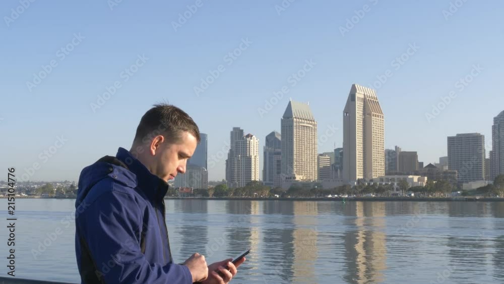 Man sms texting using app on smart phone at bacground downtown San Diego. Young man sends a sms text message on phone. Young man texting on smartphone standing on City. Traveler writes a sms message. Man sms texting using app on smart phone at bacground downtown San Diego. Young man sends a sms text message on phone. Young man texting on smartphone standing on City. Traveler writes a sms message.
