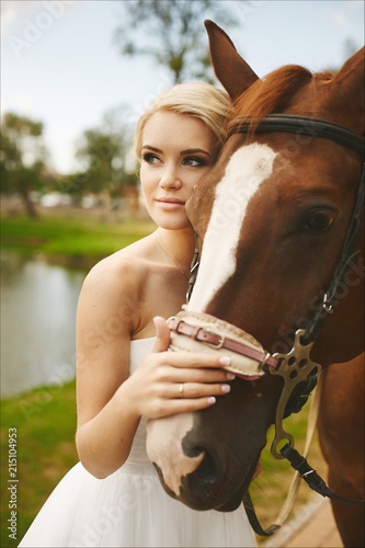 Beautiful and fashionable blonde model girl with blue eyes, with stylish hairstyle and bright makeup, in white dress posing with brown horse outdoors