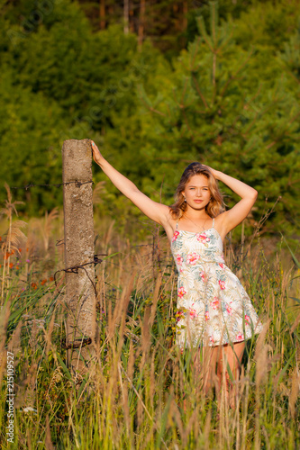 Beautiful young woman standing in a field near a concrete pillar, green grass and flowers. Outdoors Enjoy nature. Healthy smiling girl standing in tall grass