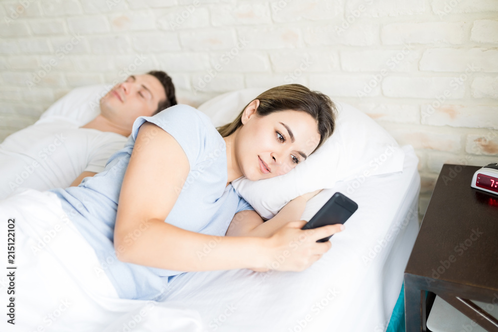 Woman Secretly Using Smartphone While Lying By Boyfriend On Bed Woman Secretly Using Smartphone While Lying By Boyfriend On Bed
