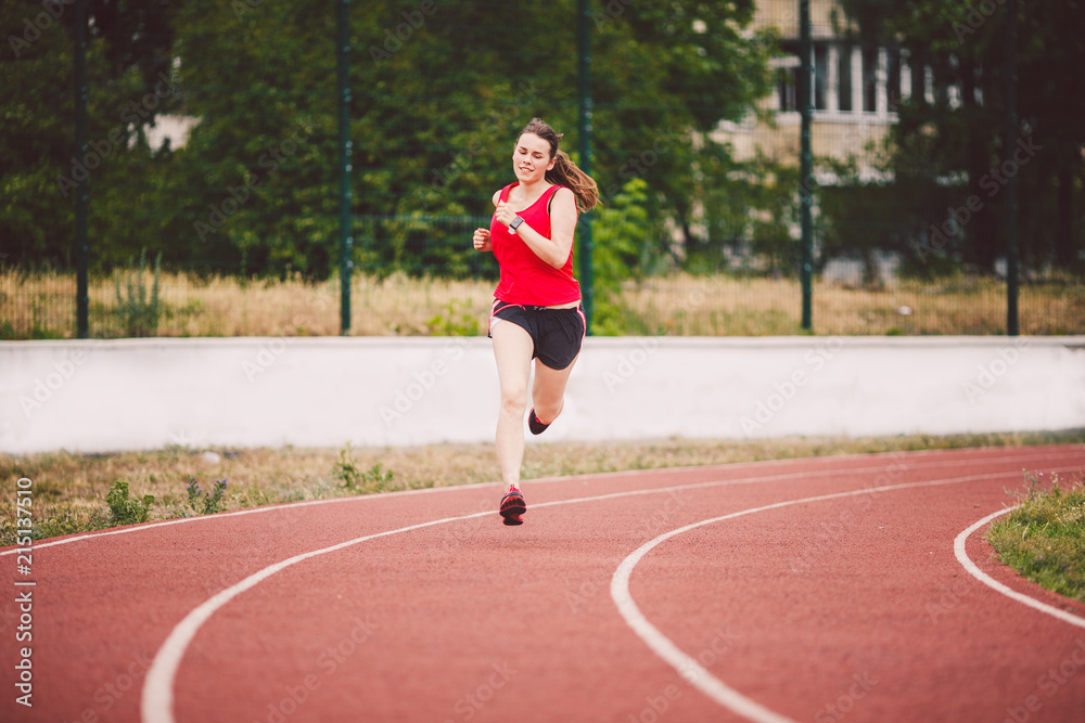 Beautiful young athlete Caucasian woman with big breasts in red T-shirt and  short shorts jogging, running in the stadium with red rubber coating Stock  Photo