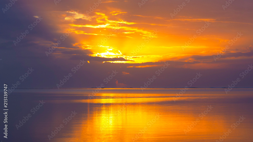 Orange tropical sun hides behind the dense clouds above the tranquil ocean.