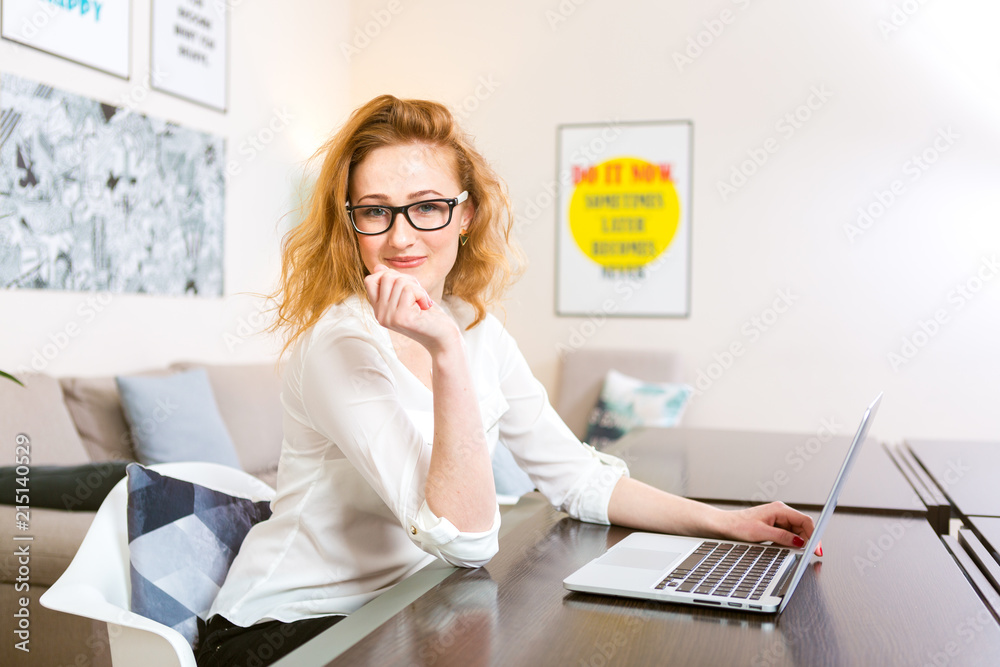 Obraz premium young woman with long red hair in white shirt and glasses for vision works, looks at the camera, uses gray laptop sitting at wooden table in a light interior. Subject business, people and technology