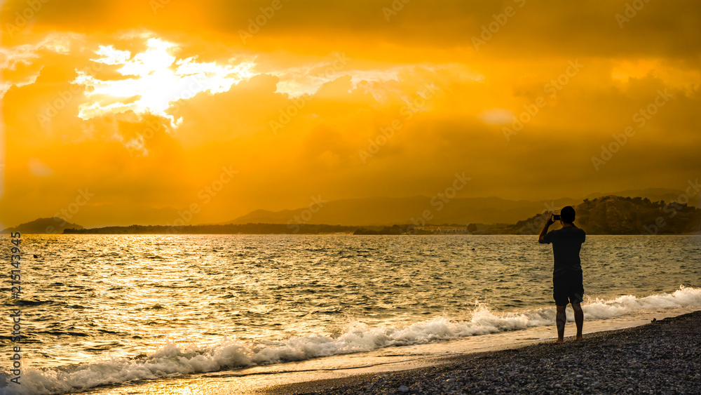 Man silhouette capturing the dramatic sunset on the beach. Beautiful blazing sunset landscape at blue sea and orange sky above it with awesome sun golden reflection on calm waves as a background. 