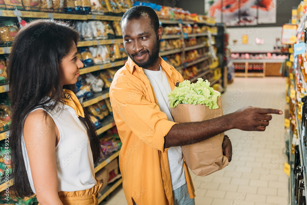 african american man with paper bag with food pointing to girlfriend in supermarket