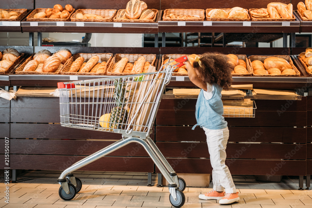 Φωτογραφία side view of little african american kid carrying shopping trolley in supermarke