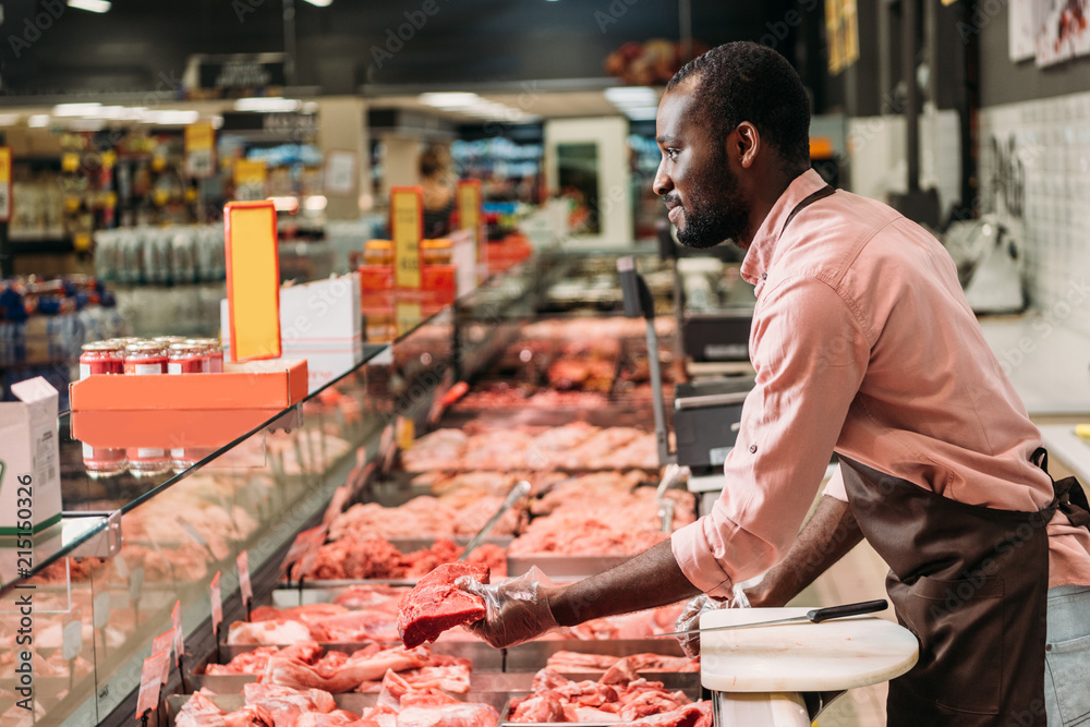 Φωτογραφία side view of african american male butcher in apron giving steak of raw meat in