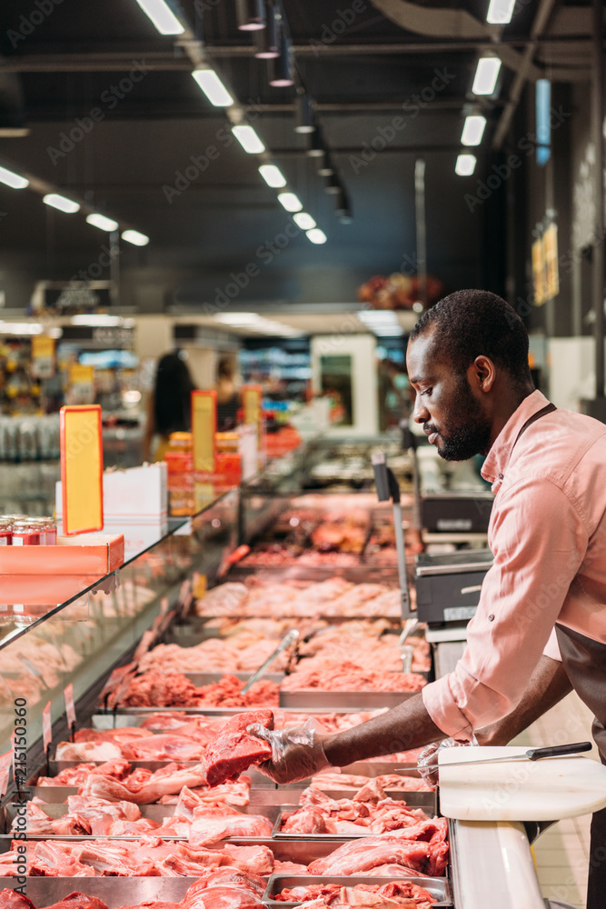 Φωτογραφία focused african american male butcher in apron taking steak of raw meat in super