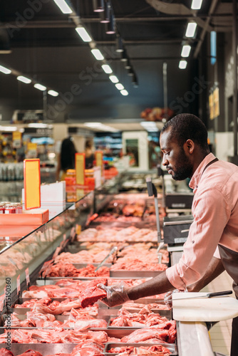 Canvastavla focused african american male butcher in apron taking steak of raw meat in super